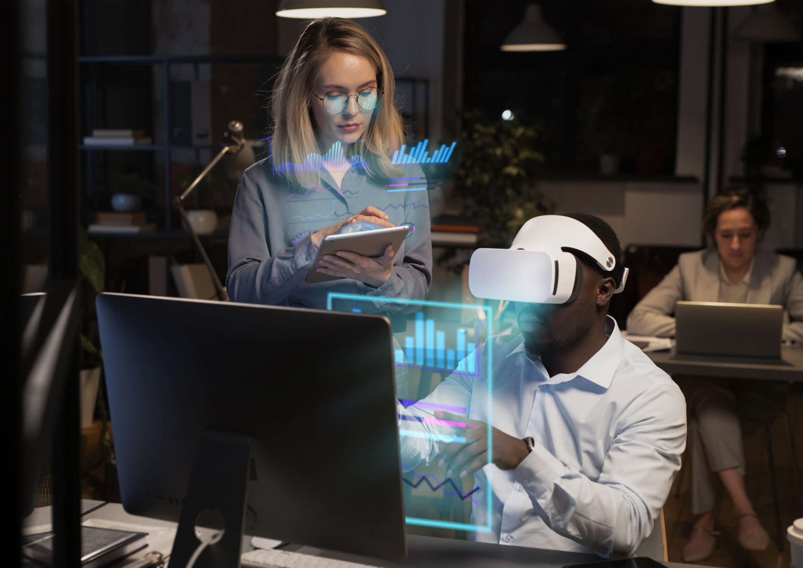 Business woman with mobile device behind a man wearing a VR headset sitting in front of a computer monitor in an office